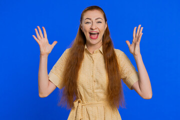 Happy joyful woman shouting, raising hands in gesture I did it, celebrating success, winning, birthday, lottery jackpot goal achievemen, good news. Young redhead girl on blue studio wall background