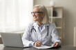© fizkes - Happy thoughtful mature professional doctor woman sitting at workplace table with laptop and documents, thinking over medical report, looking at window away, laughing, smiling