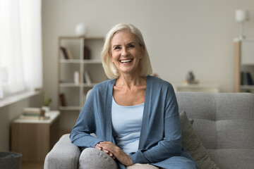 happy pretty senior woman sitting on couch at cozy home, looking at camera with toothy smile. positi