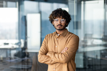 Portrait of serious thinking businessman inside office, man with crossed arms looking at camera in concentration, hispanic man at workplace wearing glasses and shirt.