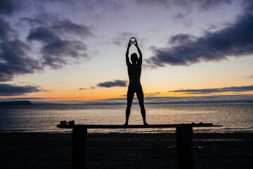 Silhouette of young woman doing workout on a bench with crystal ball(s), in front of ocean at sunrise or sunset