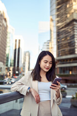 Wall Mural - Young pretty Asian business woman office professional with cellphone in hand standing on big city street holding mobile device, using smartphone, looking at cell phone, texting messages, vertical.