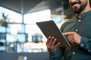 Smiling young business man holding pad computer in hands at work. Male professional employee using digital tablet fintech device standing in office checking financial market data. Close up, copy space
