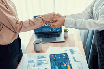 Two Asian businesswomen shaking hands in modern office after successfully analyzing piles of dashboard data papers, as importance of teamwork and integrity in the workplace concept. Enthusiastic