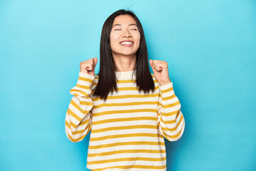 Wall Mural - Asian woman in striped yellow sweater, celebrating a victory, passion and enthusiasm, happy expression.