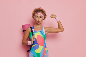 Wall Mural - Close up shot of young woman dressed in gymnastic sportswear with yoga mat poses on pink background with one fist up, barbie fashion concept, copy space