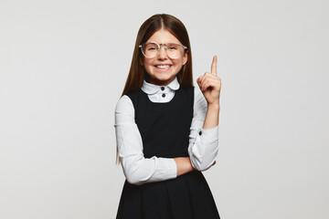 Wall Mural - Excited cute genius girl wearing glasses and school uniform smiling and pointing up with index finger against white background