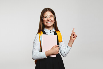 Clever little nerdy girl kid in school uniform and eyeglasses holding books and pointing up, standing isolated over white background