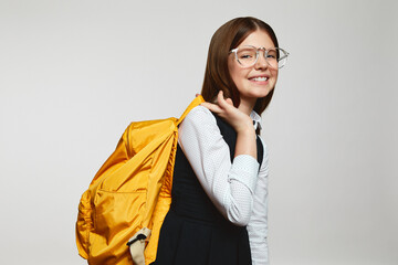 Wall Mural - Side view of cute blonde schoolgirl with glasses and uniform carrying yellow school bag and looking at camera against white background