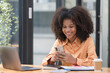 © Songsak C - Portrait of a beautiful confident businesswoman using a laptop computer holding a mobile phone sitting in a modern office. Smiling African American freelancer working online from home.