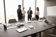 Business team of four discussing work project in office conference room, talking, standing in background at large window, white board, meeting table with stationery, notebooks, empty chairs