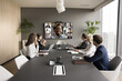 © fizkes - Young African American business leader holding video conference, talking to team online and offline. Workgroup sitting at meeting table, looking at large screen with headshots