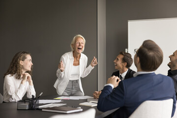 Canvas Print - Cheerful senior business leader woman and team of excited younger employees meeting at table, brainstorming on project, discussing creative ideas, laughing out loud, having fun