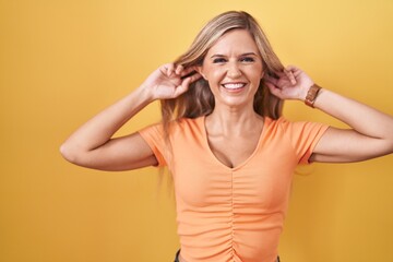 Canvas Print - Young woman standing over yellow background smiling pulling ears with fingers, funny gesture. audition problem