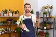 Middle age man with beard working at florist shop showing smartphone screen making fish face with mouth and squinting eyes, crazy and comical.