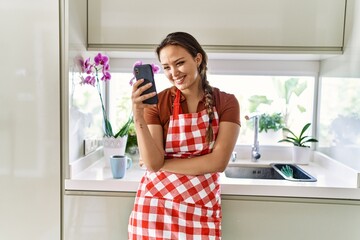 Poster - Young beautiful hispanic woman wearing apron using smartphone at the kitchen