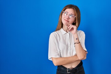 Wall Mural - Beautiful woman standing over blue background with hand on chin thinking about question, pensive expression. smiling with thoughtful face. doubt concept.