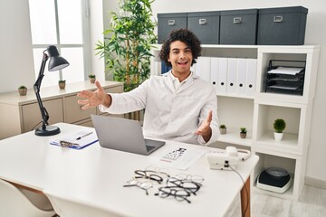 Canvas Print - Hispanic man with curly hair working at optician office looking at the camera smiling with open arms for hug. cheerful expression embracing happiness.