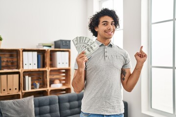 Canvas Print - Hispanic man with curly hair holding 100 dollars banknotes smiling happy pointing with hand and finger to the side