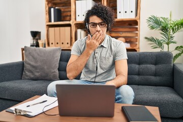 Wall Mural - Hispanic man with curly hair doing online session at consultation office covering mouth with hand, shocked and afraid for mistake. surprised expression