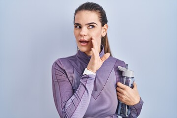 Poster - Beautiful woman wearing sportswear holding water bottle hand on mouth telling secret rumor, whispering malicious talk conversation