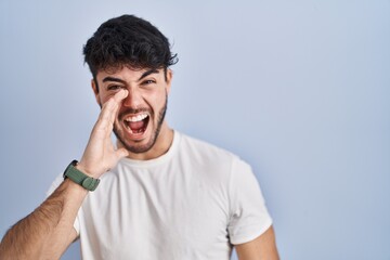 Wall Mural - Hispanic man with beard standing over white background shouting and screaming loud to side with hand on mouth. communication concept.