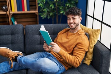 Young hispanic man reading book sitting on sofa at home