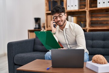 Poster - Young hispanic man psychologist talking on smartphone holding clipboard psychology center