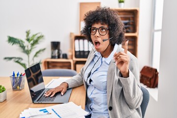 Poster - Black woman with curly hair wearing call center agent headset at the office amazed and surprised looking up and pointing with fingers and raised arms.