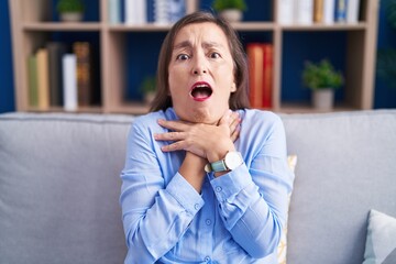 Canvas Print - Middle age hispanic woman sitting on the sofa at home shouting and suffocate because painful strangle. health problem. asphyxiate and suicide concept.