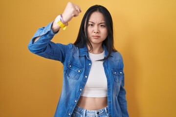 Sticker - Young asian woman standing over yellow background angry and mad raising fist frustrated and furious while shouting with anger. rage and aggressive concept.