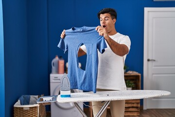 Sticker - Hispanic man ironing holding burned iron shirt at laundry room afraid and shocked with surprise and amazed expression, fear and excited face.