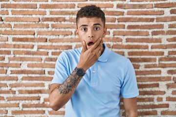 Canvas Print - Brazilian young man standing over brick wall looking fascinated with disbelief, surprise and amazed expression with hands on chin