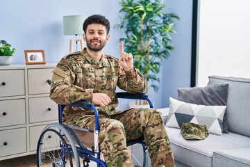 Poster - Arab man wearing camouflage army uniform sitting on wheelchair showing and pointing up with finger number one while smiling confident and happy.