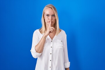 Sticker - Young caucasian woman standing over blue background asking to be quiet with finger on lips. silence and secret concept.