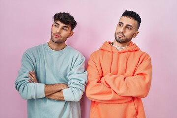 Wall Mural - Young hispanic gay couple standing over pink background looking to the side with arms crossed convinced and confident
