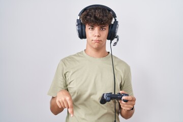 Hispanic teenager playing video game holding controller pointing down looking sad and upset, indicating direction with fingers, unhappy and depressed.