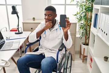 Wall Mural - African american doctor man sitting on wheelchair holding smartphone serious face thinking about question with hand on chin, thoughtful about confusing idea