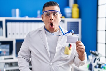 Canvas Print - Hispanic young man working at scientist laboratory holding id card scared and amazed with open mouth for surprise, disbelief face