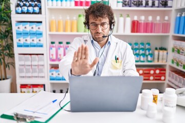 Sticker - Hispanic young man working at pharmacy drugstore working with laptop with open hand doing stop sign with serious and confident expression, defense gesture