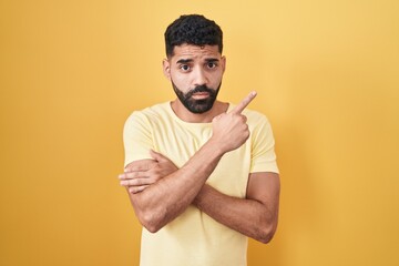 Wall Mural - Hispanic man with beard standing over yellow background pointing with hand finger to the side showing advertisement, serious and calm face