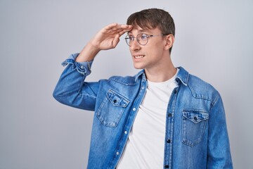 Poster - Caucasian blond man standing wearing glasses very happy and smiling looking far away with hand over head. searching concept.