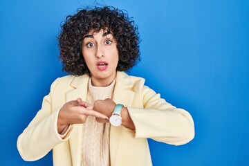 Poster - Young brunette woman with curly hair standing over blue background in hurry pointing to watch time, impatience, upset and angry for deadline delay