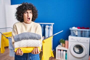 Wall Mural - Young brunette woman with curly hair holding clean laundry angry and mad screaming frustrated and furious, shouting with anger. rage and aggressive concept.