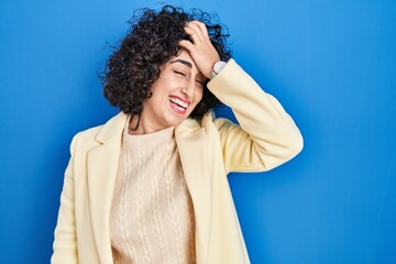 Sticker - Young brunette woman with curly hair standing over blue background surprised with hand on head for mistake, remember error. forgot, bad memory concept.