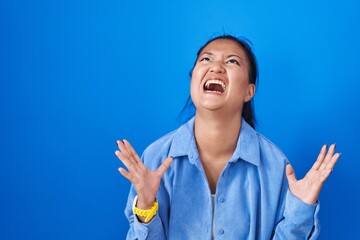 Poster - Asian young woman standing over blue background crazy and mad shouting and yelling with aggressive expression and arms raised. frustration concept.