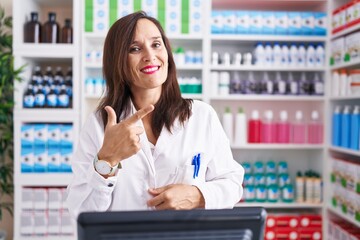 Poster - Middle age brunette woman working at pharmacy drugstore cheerful with a smile of face pointing with hand and finger up to the side with happy and natural expression on face