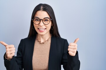 Poster - Young brunette woman standing over blue background success sign doing positive gesture with hand, thumbs up smiling and happy. cheerful expression and winner gesture.