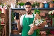 Hispanic young man working at florist shop showing smartphone screen in shock face, looking skeptical and sarcastic, surprised with open mouth