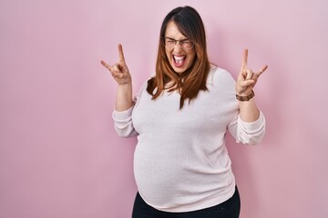 Poster - Pregnant woman standing over pink background shouting with crazy expression doing rock symbol with hands up. music star. heavy music concept.